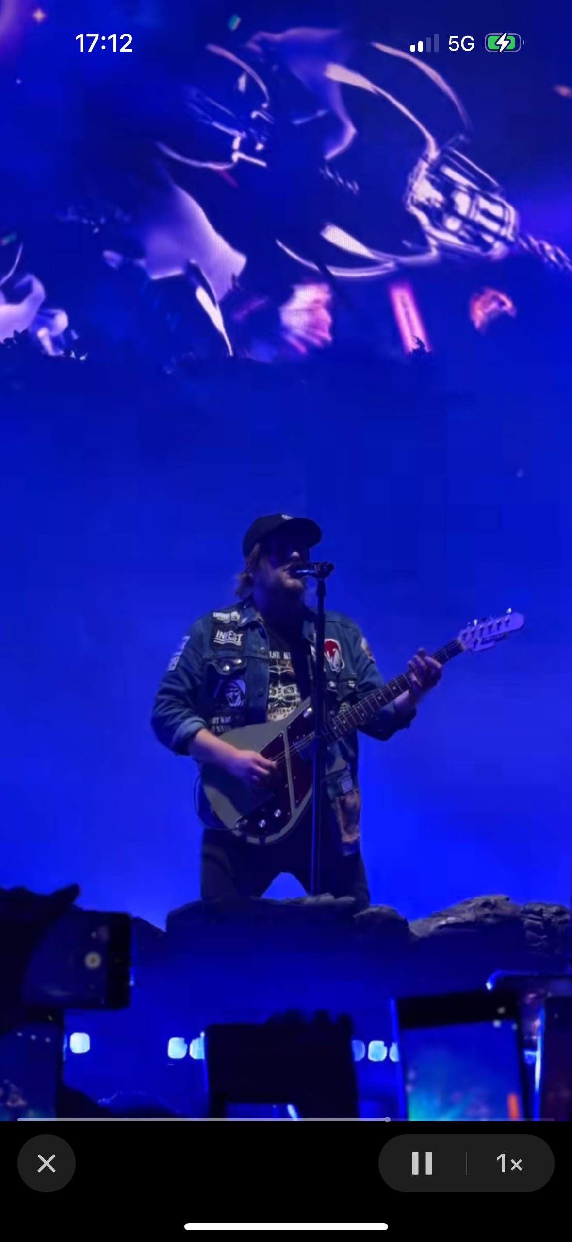 a musician is performing on stage at a concert. he is wearing a black jacket and a black hat. he is playing a guitar and singing into a microphone. the stage is lit up with blue lights and the crowd is in the background.  the crowd is blurry and dark. there are other lights and speaker equipment in the background, blurry and dark, behind the musician.  a microphone stand can be seen next to the musician.