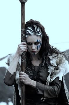 a fierce female warrior stands in a cloudy, wild landscape, holding a spear and staring intently.  she is wearing a fur garment and leather armor and is painted with tribal markings. she appears to be ready to fight, fierce and strong.
