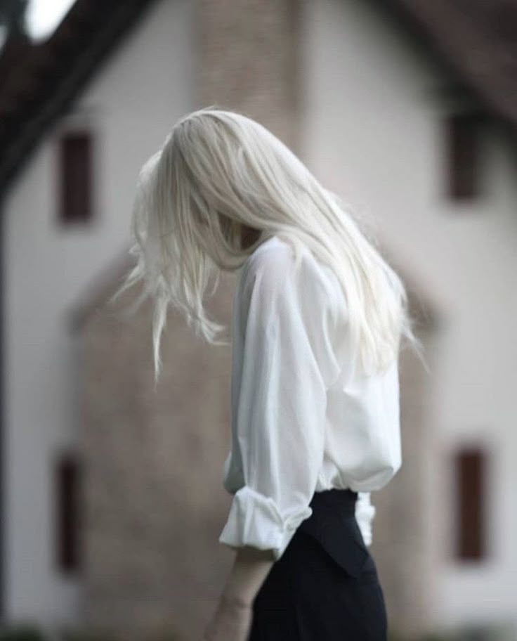 a young woman with long, platinum blonde hair is standing in front of a blurred background of a house. she is wearing a loose white shirt and a black skirt. her hair is hanging down, partially obscuring her face. the overall image is monochromatic and has a soft, aesthetic feel.