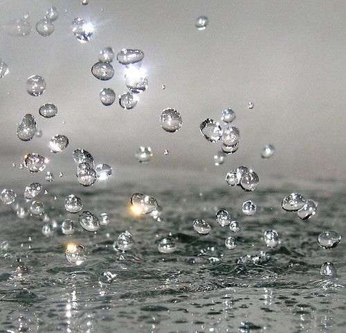 the image shows numerous water droplets suspended in the air above a wet surface. the droplets vary in size and appear to be caught midair, with some reflecting light creating bright highlights. the wet surface below is textured and also reflects light. the background is a blurred light gray.