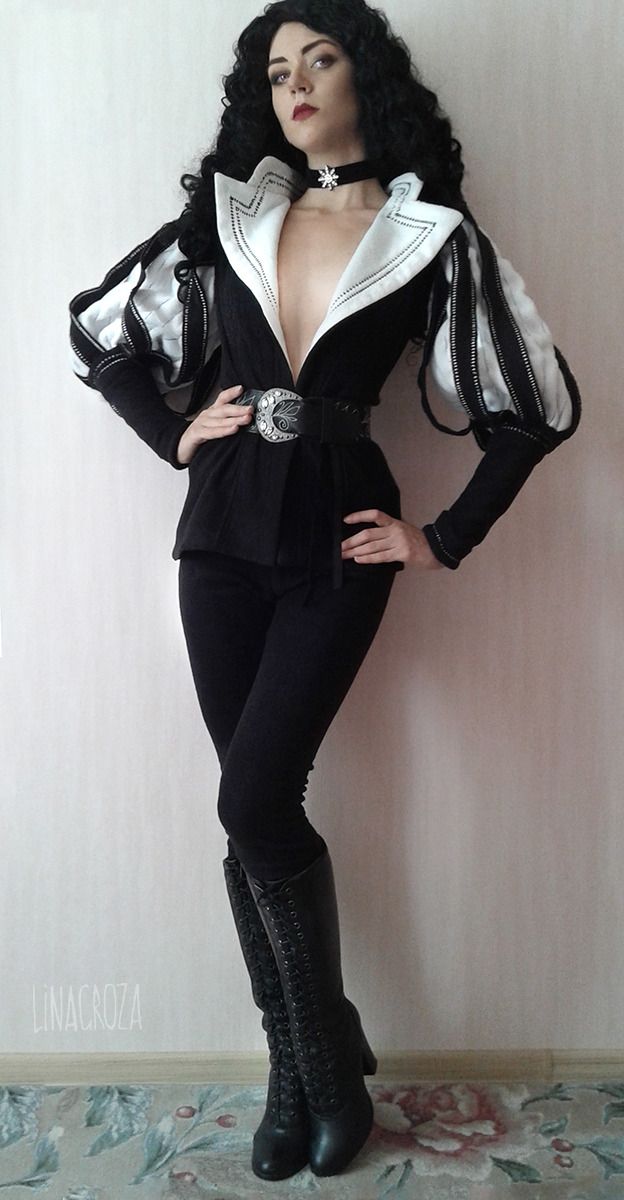 a woman is cosplaying as triss merigold from the witcher 3 video game. she's wearing a black and white costume with puffed sleeves, a black belt with a silver buckle, and black laceup boots.  she has long dark hair and is posing against a lightcolored wall with a patterned carpet on the floor. the overall style is dark and elegant, reflective of the character's appearance in the game.