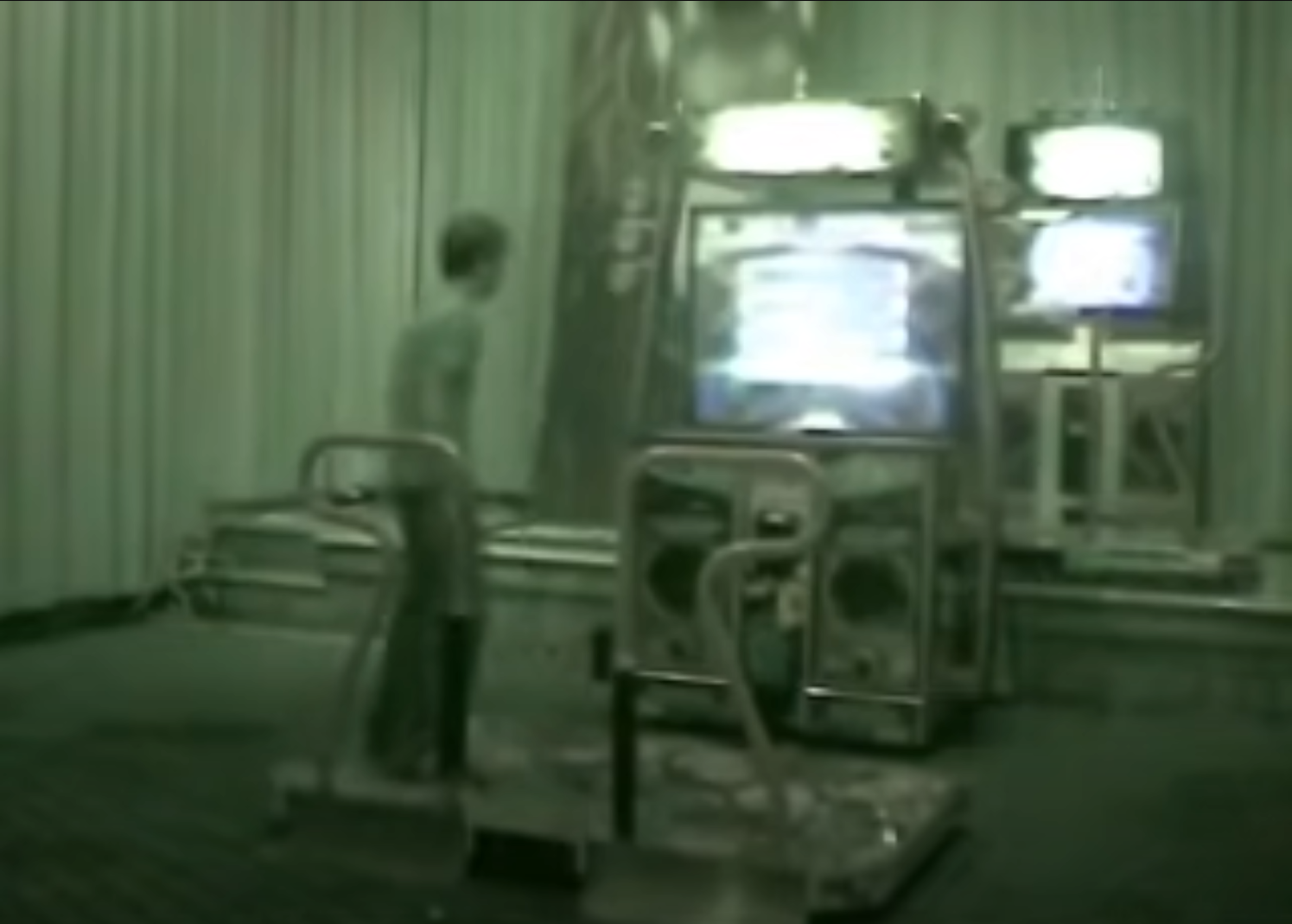 a man is playing an arcade racing game inside what appears to be a mostly empty arcade. the image has a green tint and the lighting is dim.