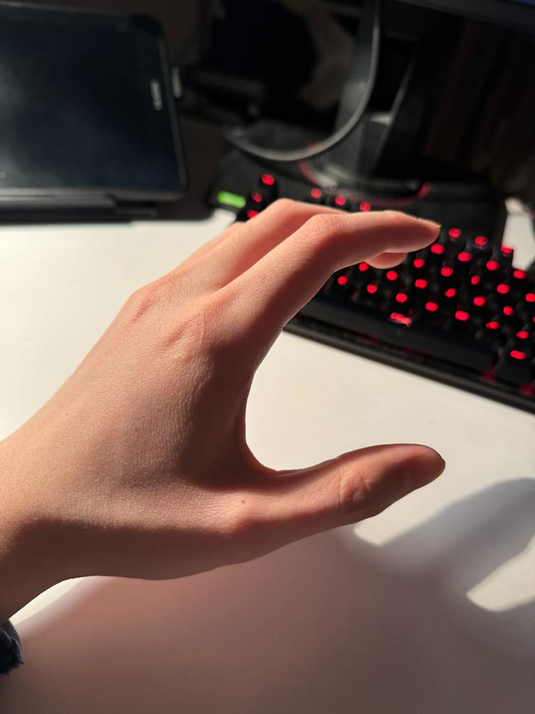 fairskinned hand is extended, fingers curved inward in a grabbing or reaching gesture. the hand is positioned above a white surface with a red and black keyboard in the background, lit by natural light creating a strong shadow on the surface.