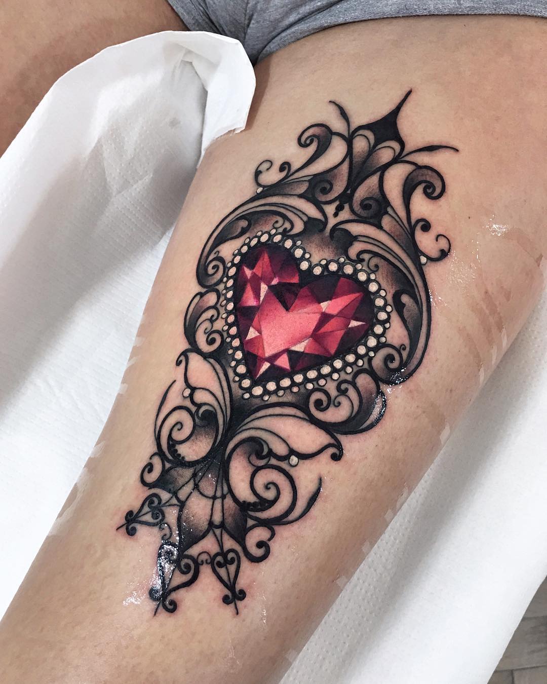 a fresh tattoo of an ornate heartshaped diamond within a filigree design on a woman's upper thigh. the tattoo is primarily black and gray with red accents in the diamond. the photo is taken from above looking down at the tattoo.