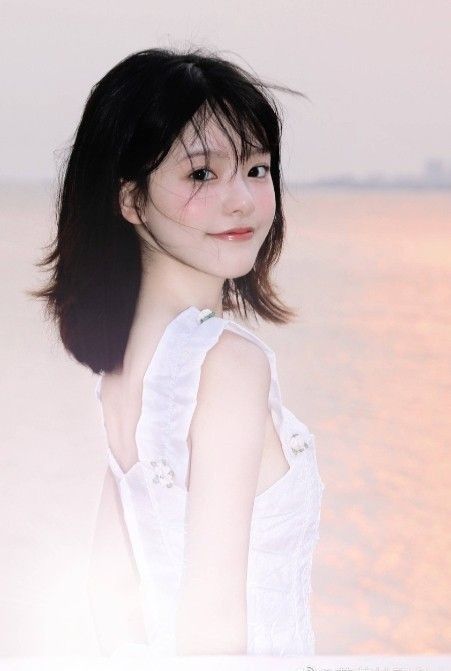 a young woman with short black hair is standing on a beach with the setting sun in the background. she is wearing a white dress with ruffles and smiling slightly. the image is taken from a side view and the lighting is soft and natural.