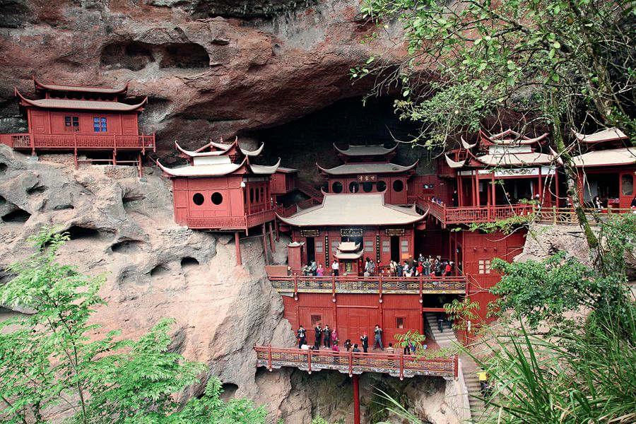 a wide, full shot depicts a red, wooden, multitiered architectural complex built into the side of a rocky cliff or cave. the structure resembles a temple or monastery, with traditional asian architectural elements such as curved roofs and ornate details. many people are visible, exploring the complex and gathering on platforms. lush green vegetation surrounds the structure, softening the harshness of the rock. the natural rock formation with many caves and hollows contrasts with the constructed building.