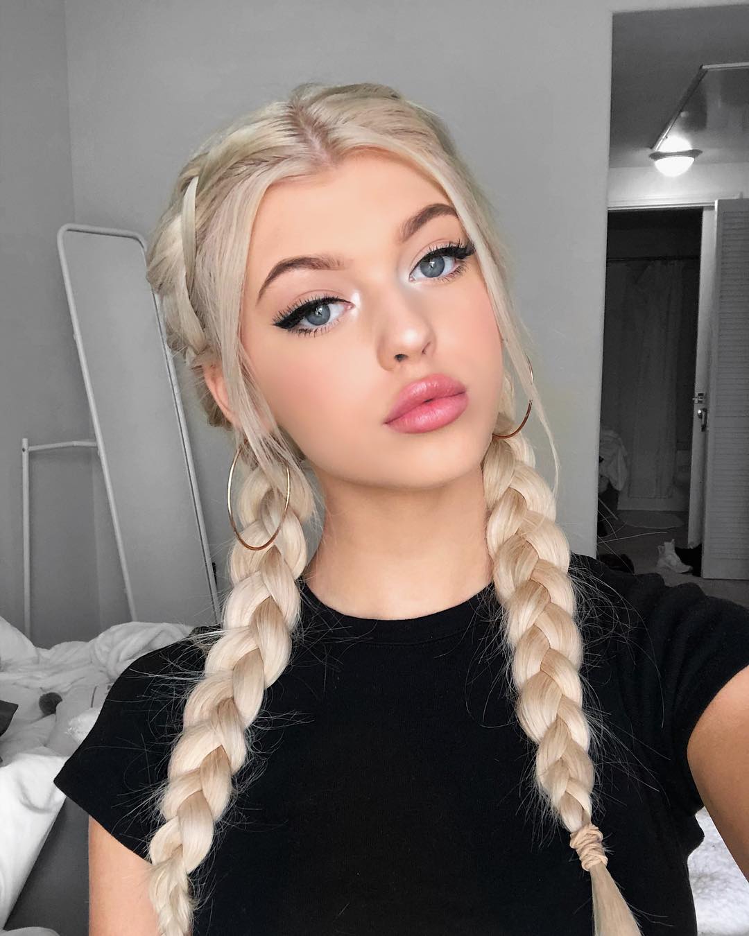 a young woman with blonde hair styled in two braids, wearing large hoop earrings and a black tshirt, takes a selfie in what appears to be her bedroom.