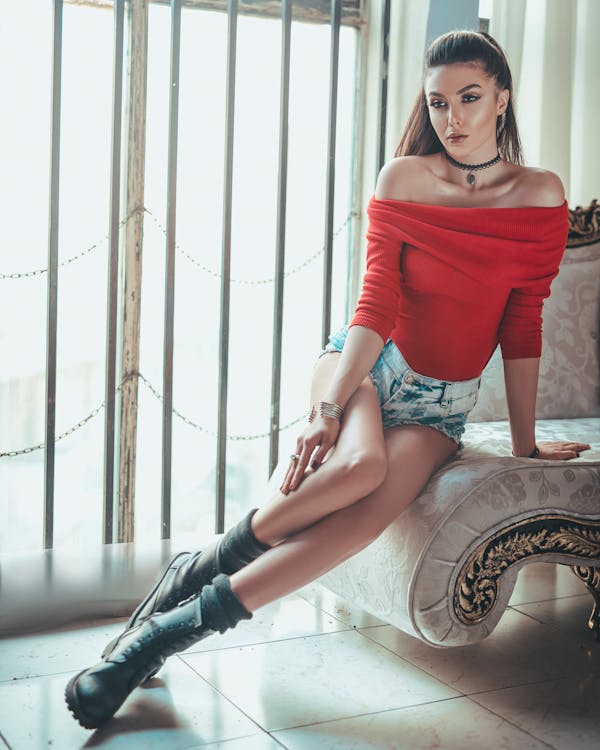 a young, attractive woman with long dark brown hair is sitting on an ornate chaise lounge. she's wearing a red offtheshoulder top, short denim shorts, and stylish combat boots.  she's accessorized with a necklace and bracelets. the setting is indoors, near a window with metal bars, creating a somewhat moody yet fashionable atmosphere.  she is posing confidently, showcasing her attire and looks directly at the camera. the overall image is stylish and suggests a fashion or editorial shoot.