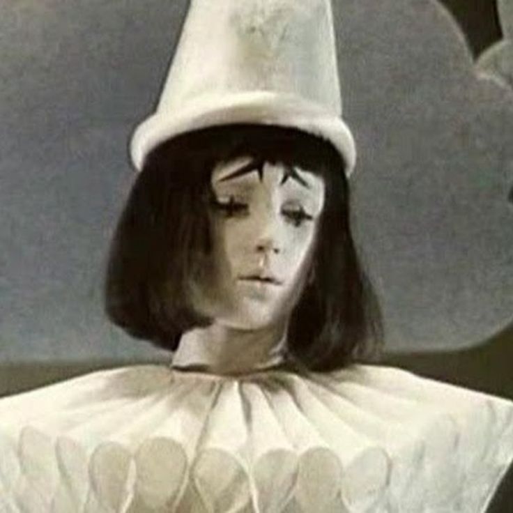 a sad clown girl with a white hat sits and looks down. the image is a closeup and focuses on the clown's face and the top of her costume. the background is a grey and white wall.