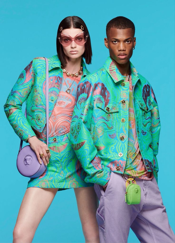 a fashion photograph showcasing a male and female model dressed in matching versace clothing. both models wear a vibrant teal jacket and matching bottoms. the woman pairs her outfit with pastel purple sunglasses and a matching bag. the man wears lavender pants. the clothing features a psychedelic, swirling print. the backdrop is a solid teal color. the image is professionally shot and emphasizes the highfashion aspect of the clothing.