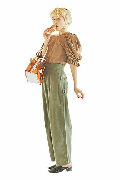 a young blonde woman is posing in a brown puffsleeved shirt, green pants, black shoes, and holding a brown snack while carrying a transparent and white bag.  she is standing with a slight smile, facing the camera.