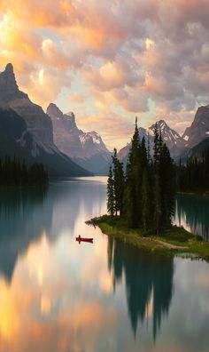 a single red canoe floats on a glassy lake, mirroring the majestic mountains and a small island with tall evergreen trees. the scene is bathed in the warm golden light of a sunrise, painting the sky with hues of orange and pink.  this serene landscape evokes a sense of peace and tranquility.