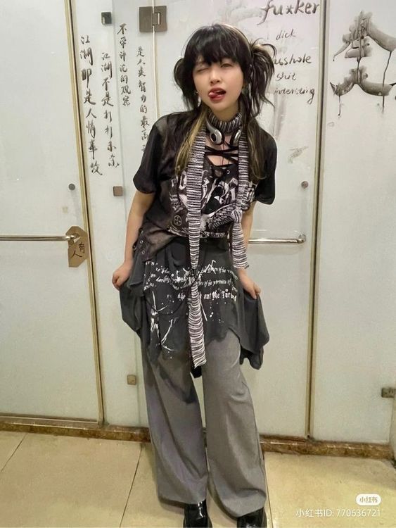 young asian woman with dark hair styled in a bob with side bangs, is standing in a restroom. she's wearing a black graphic tshirt with a grey, asymmetrical tunic or oversized shirt tied at the waist, and loosefitting grey trousers.  a grey and black striped scarf is loosely draped around her neck. the wall behind her is white and features graffiti including the word "fuxker" and chinese characters.  she has a slightly mischievous or playful expression.  the overall style is edgy and alternative.