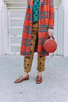 a woman wearing a red plaid coat, brown polka dot pants, brown shoes, and a brown handbag walks down the street in an urban environment. she is dressed in a casual chic style.