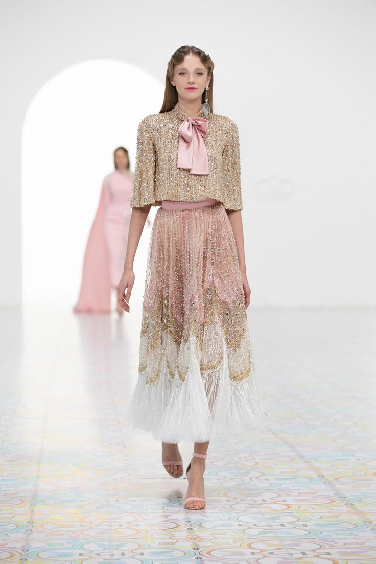 the image shows a fashion model walking on a runway during a fashion show.  she is wearing a stunning, intricately designed gown. the dress features a short, sequined, goldtoned top with a pink bow at the neckline. the skirt is a longer piece, made from a delicate tulle material, and is ombrecolored, progressing from pale pink to gold and finishing with a white fringe. the model's makeup and hairstyle are simple and elegant, complementing the ethereal quality of the dress.  another model wearing a pink gown is visible in the background, suggesting a fashion show setting. the floor is patterned with colorful, soft tones. the overall feel is one of luxury, high fashion, and refinement.