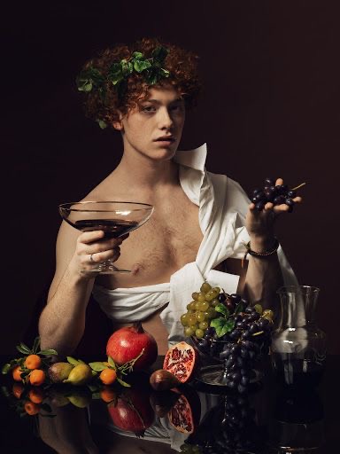 the image depicts a young man posed as bacchus, the roman god of wine and revelry. he is positioned at a table, shirtless with an unbuttoned white shirt draped around his torso. he wears a crown of ivy leaves on his head. in his right hand, he holds aloft a glass of red wine, and in his left, a cluster of grapes. the table before him is laden with a bountiful still life of fruits, including grapes, pomegranates, figs, and oranges. a crystal decanter of red wine also sits on the table. the composition is reminiscent of classical renaissance portraiture, with dramatic chiaroscuro lighting highlighting the subject and creating a sense of theatricality. the dark background further emphasizes the subject and the richness of the scene.  the image evokes a sense of opulence, pleasure, and celebration, aligning with the themes associated with the god bacchus.