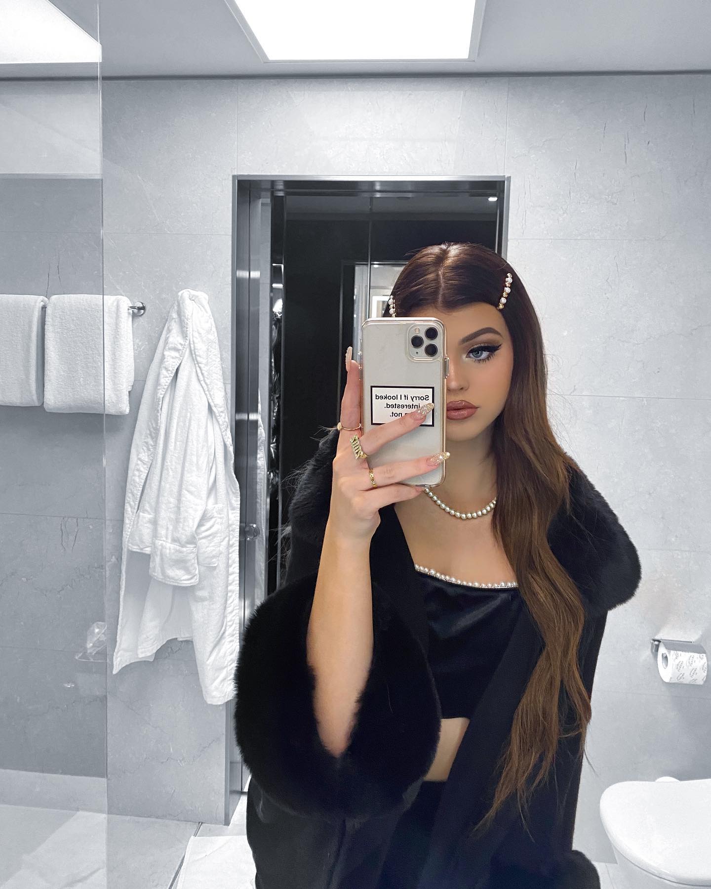 a woman is taking a selfie in a bathroom mirror. she's wearing a black top, a pearl necklace, and has hair clips in her hair. in the background, there's a white robe hanging on the wall, and towels.