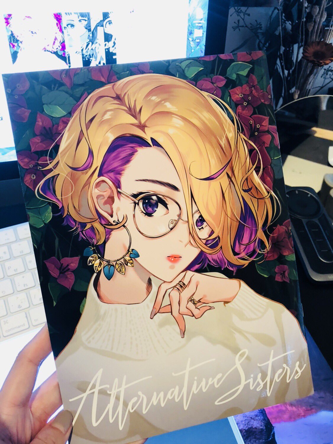 the image shows a printed artbook cover featuring an animestyle illustration of a girl with yellow and purple hair. she wears round glasses and has a delicate earring. the background features stylized flowers in shades of pink and purple. the title "alternative sisters" is written in cursive script at the bottom of the cover. the photo is taken indoors with artificial lighting, capturing the artbook being held by a hand.