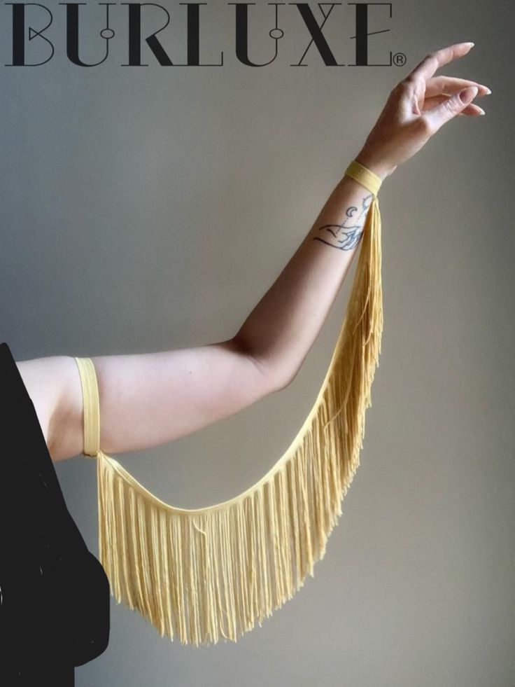 an image of a person's arm with yellow fringe jewelry, showing the wrist, the arm and the hand. on the arm is a tattoo. the upper left side of the image has the word "burluxe" with a registered trademark symbol.