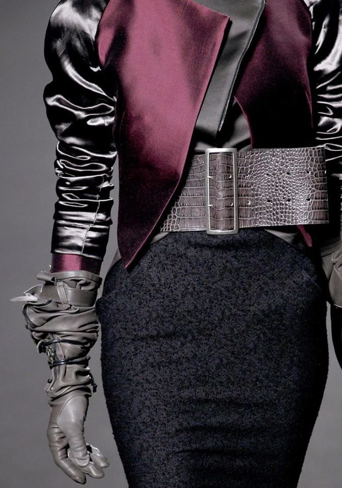a woman is posing in a studio setting, she's wearing a black and burgundy outfit that includes a jacket, a skirt, gloves and a belt. she is looking very chic and elegant.
