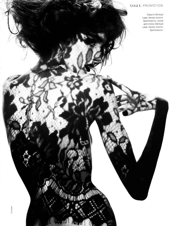 a black and white, highfashion photograph of a woman, possibly a model, posing with her back to the camera.  she is wearing a black lace garment, which casts shadows that create a lacelike pattern on her skin. her hair is styled up, and the overall aesthetic is sensual, dark, and elegant. the image is reminiscent of classic high fashion photography, with a strong emphasis on texture and shadow play. the background is plain white, drawing all attention to the model and the intricate detail of the lace.  vogue branding appears in the upper right corner.