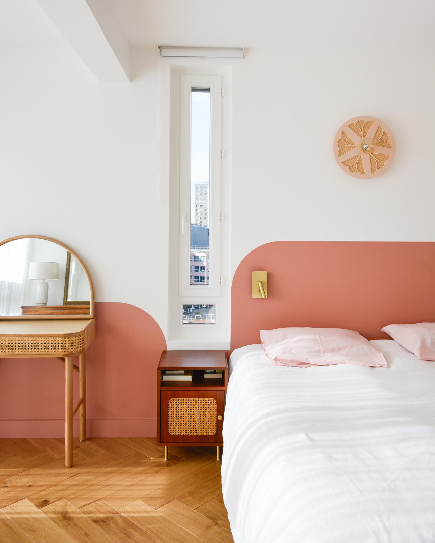 a bedroom is shown with a bed, nightstand, vanity, and mirror. the walls are white and pink, with a pink halfcircle pattern on the wall behind the vanity and a pink headboard behind the bed. a nightstand is beside the bed, and a vanity with a round mirror is on the opposite side. there is a small window in the wall. a piece of artwork is above the bed. the floor is wood with a herringbone pattern.