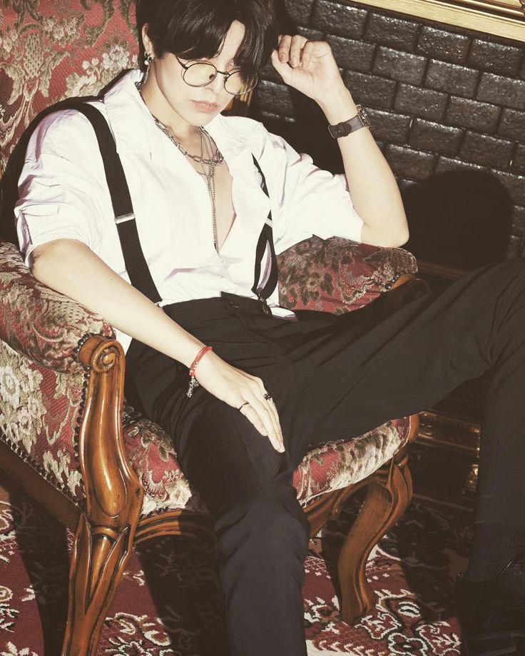 a man with black hair is sitting in a vintage chair. he is wearing a white shirt, black pants, suspenders, glasses, jewelry, and a watch. he has his hand on his leg and is looking down. the chair is red and ornate. the background is a brick wall.