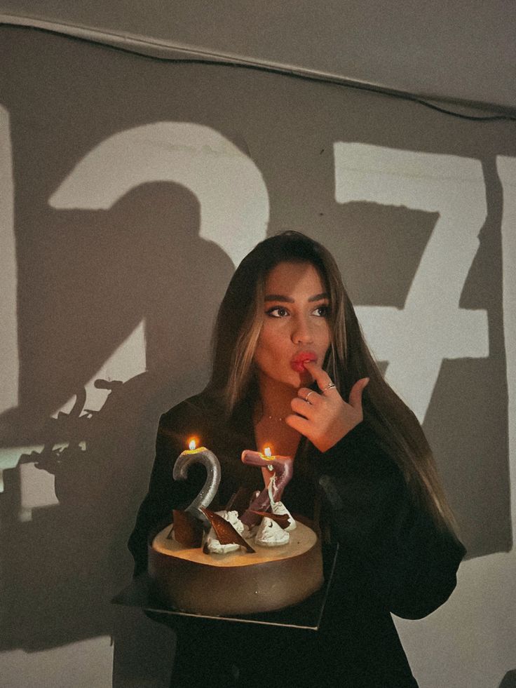 the image shows a young woman with long brown hair wearing a black blazer. she is holding a small, round birthday cake with lit "27" candles. the woman appears to be licking or tasting the frosting on her cake.  the background features a wall with the numbers "27" projected as large shadows. the overall mood is intimate and celebratory, suggestive of a birthday. the lighting is relatively dim, focusing attention on the woman and her cake.