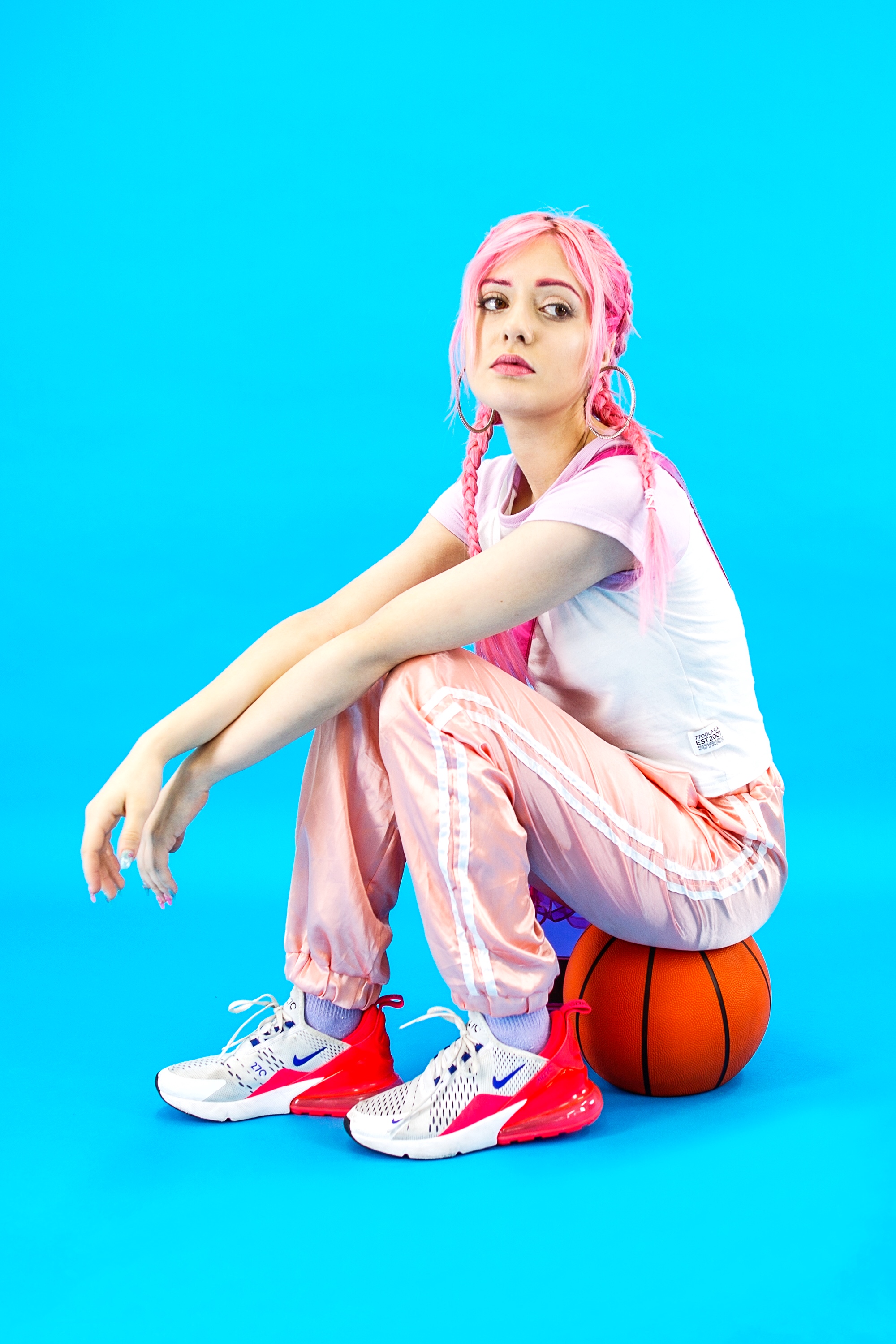 a young woman with bright pink hair is sitting on a basketball. she is wearing pink pants and a white shirt. the woman is wearing white and red nike air max 270 sneakers. the background is blue. she is looking to the side with a confident and playful expression.