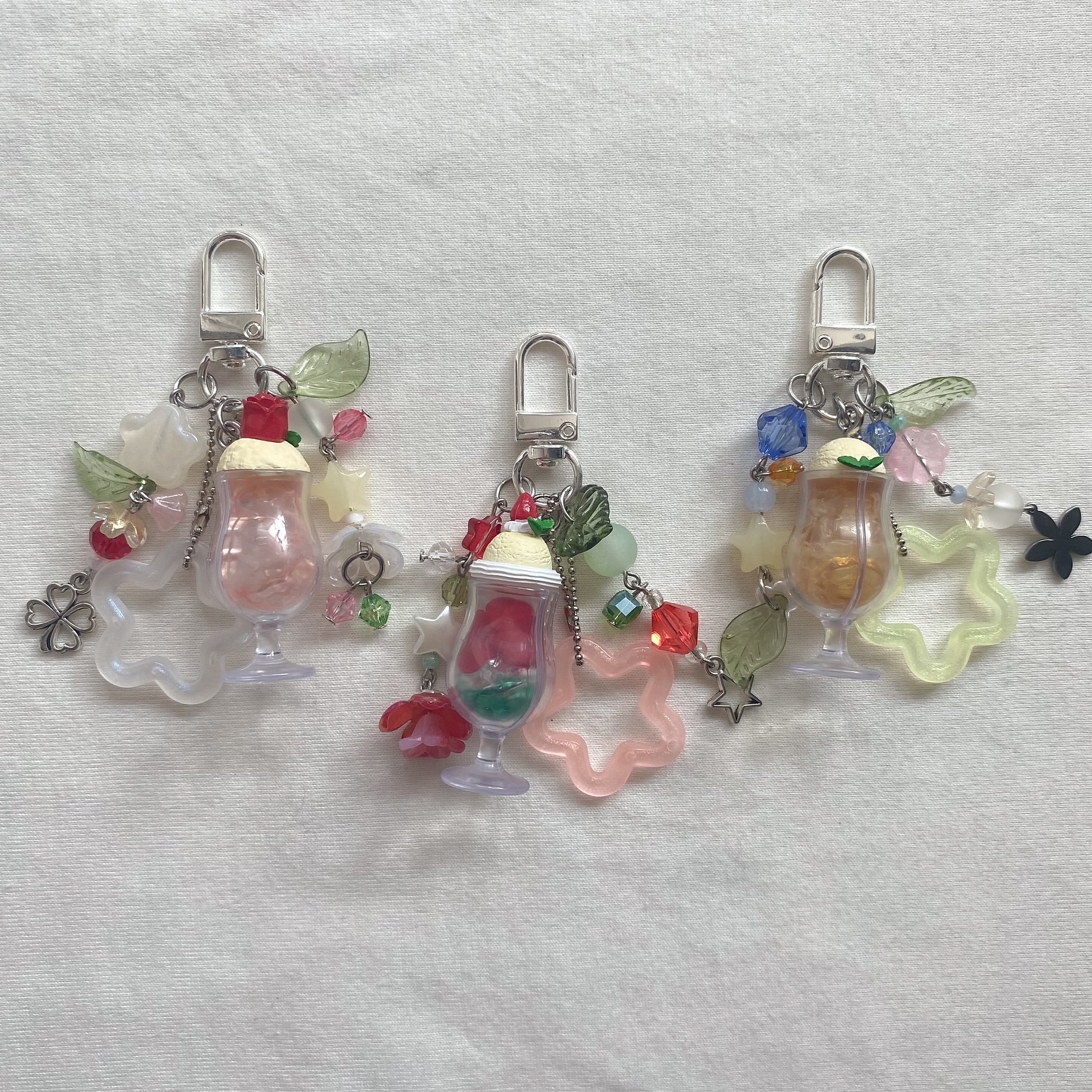the image shows three keychains with unique charms attached. each keychain features a cocktail glass charm filled with different colored liquid and topped with what appears to be a dollop of whipped cream and a cherry. the keychains are adorned with colorful beads, plastic leaves, plastic star, clover and flower charms.