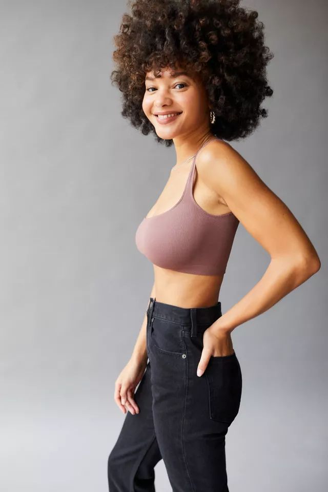 a young woman with an afro smiles at the camera. she's posing in a studio against a simple gray background. she is wearing a mauve bralette and black high waisted jeans, with her hand in her pocket. the image is welllit with soft lighting, taken from an eyelevel perspective.