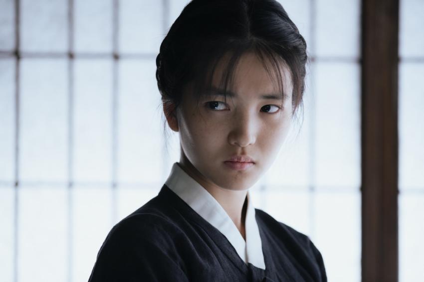 a closeup shot of a young woman wearing traditional korean clothing (hanbok). she has dark hair pulled back, a white collar, and a serious expression on her face as she looks off to the side. the background shows a gridpatterned window.