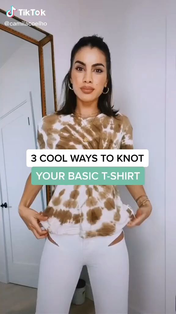 a woman in a tiedye tshirt and white jeans stands in front of a mirror and white door, demonstrating how to knot a tshirt. she is wearing hoop earrings, a gold chain, and bracelets. a tiktok logo and the username @camilacoelho are visible at the top of the image. a text overlay reads "3 cool ways to knot your basic tshirt".