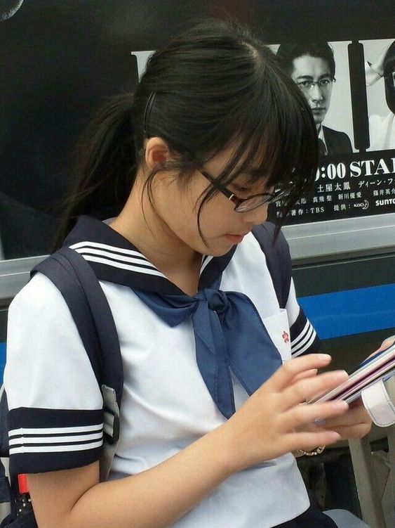 a japanese school girl in uniform is pictured reading a book on public transport, likely a train. she is wearing a white and navy sailorstyle top with a navy necktie. her dark hair is pulled back in a ponytail and she wears glasses. the background features a partially obscured advertisement. the image is naturally lit and taken from an eyelevel perspective.