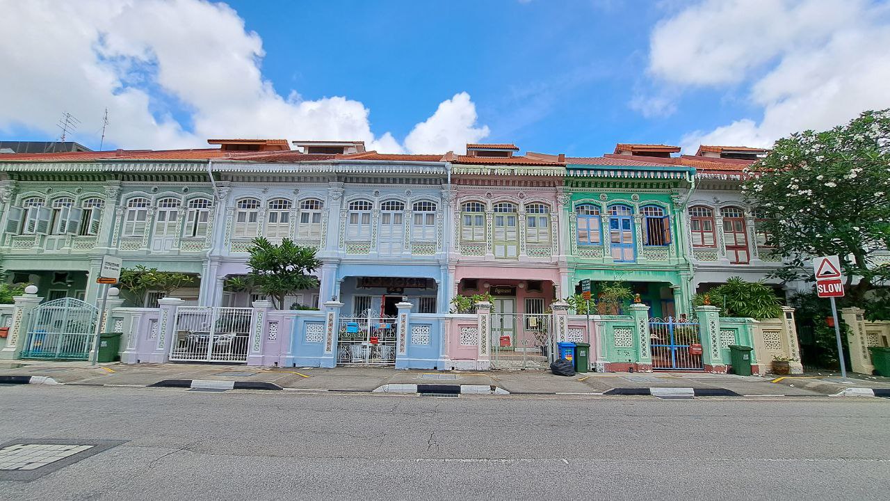 a row of colorful shop houses in singapore. the houses are all painted in different shades of green, blue, pink, and white. they have intricate details, including windows, doors, and gates. the houses are lined up along a street with a sidewalk and curb. there is a tree in front of the houses, and a sign that says "slow" is visible in the background. the sky is blue and cloudy.