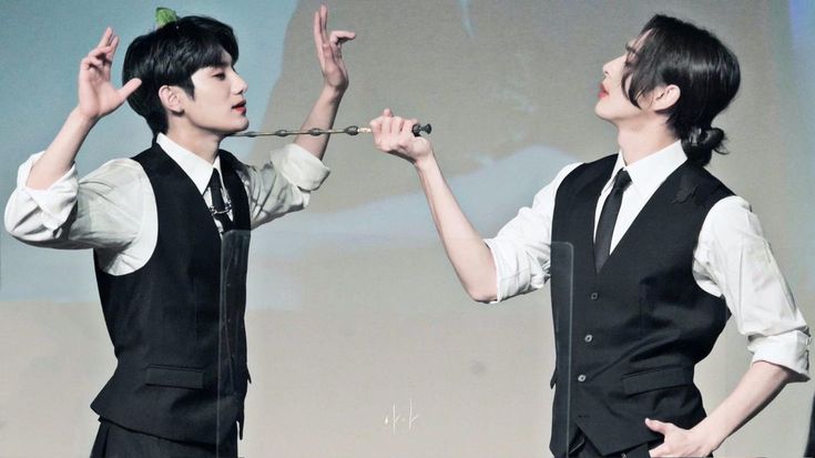 two asian men dressed in formal wear pose playfully for a photograph, mimicking a magic duel with a wand. the man on the left holds the wand, pointed at the other man, while the man on the right has his hand raised in defense. the setting appears to be indoors with bright artificial lighting.