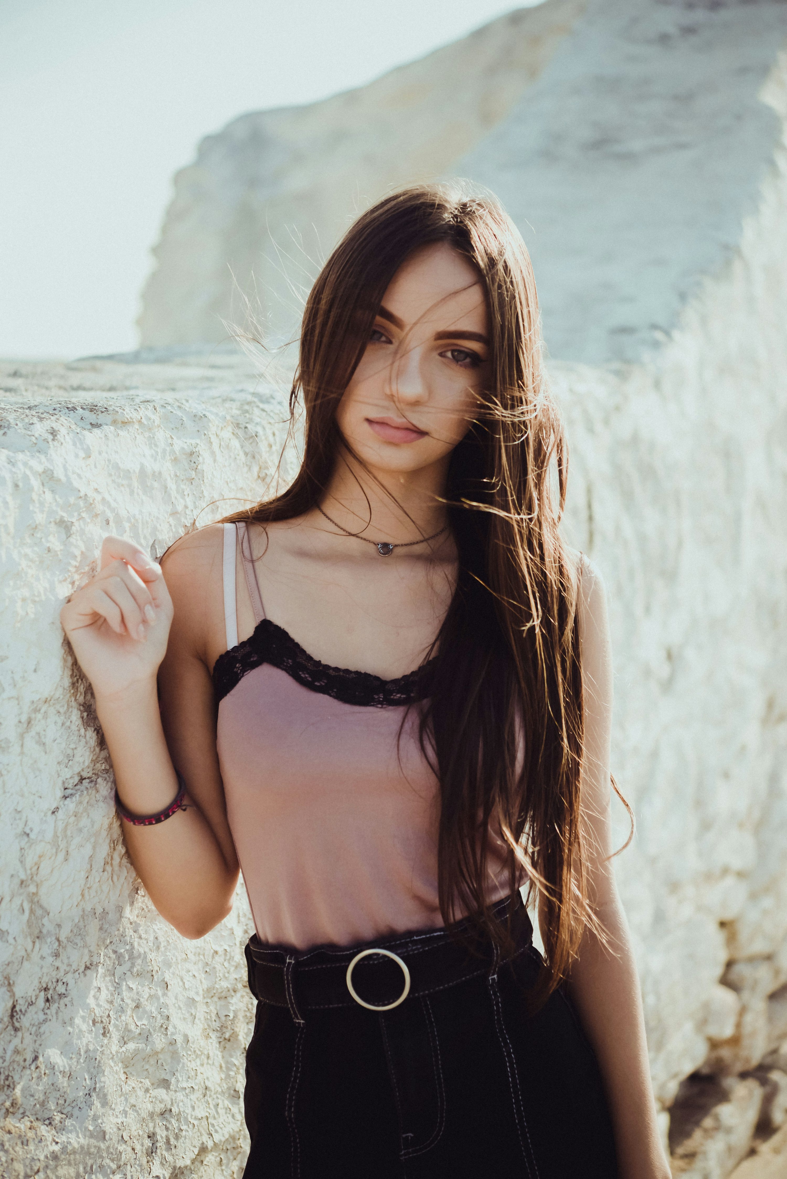 a young woman with long brown hair stands against a white rock wall. the wind is blowing her hair. she is wearing a dusty rose colored tank top with black lace trim and black pants with a belt. she has a calm and serene expression. the background is a sunny outdoor landscape with white rocks and stones.