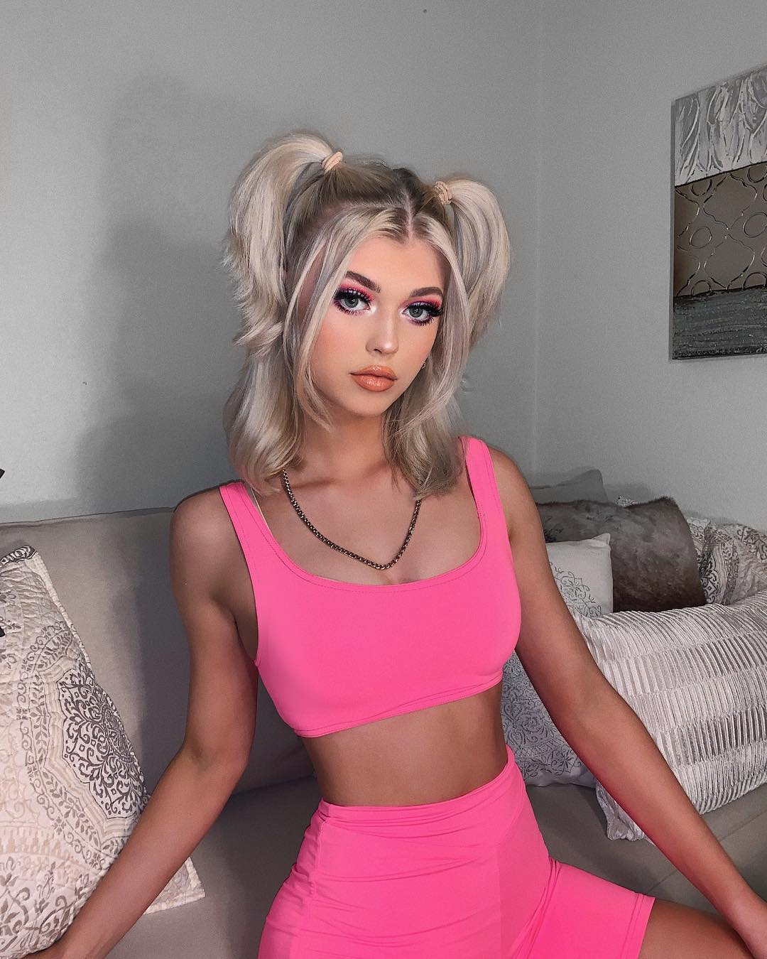 a woman with blonde hair styled in pigtails, wearing a pink top and shorts, is sitting on a couch with pillows. she has a necklace and is wearing makeup.