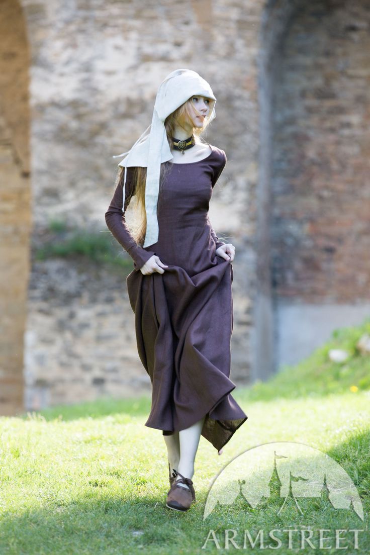a woman in a long brown medieval dress and white hood walks through a grassy field with a stone wall in the background. she is participating in a historical reenactment or festival. the woman's blonde hair is visible underneath the hood. she is wearing a simple brown dress with long sleeves, a white hood over her head, and a brown belt around her waist. her hair is visible under the hood. she appears to be moving or walking away from the camera, and her face is turned slightly towards the right side of the image. the stone wall is out of focus in the background. the grass in the foreground is in focus. the woman's dress and hood are the focal points of the image.
