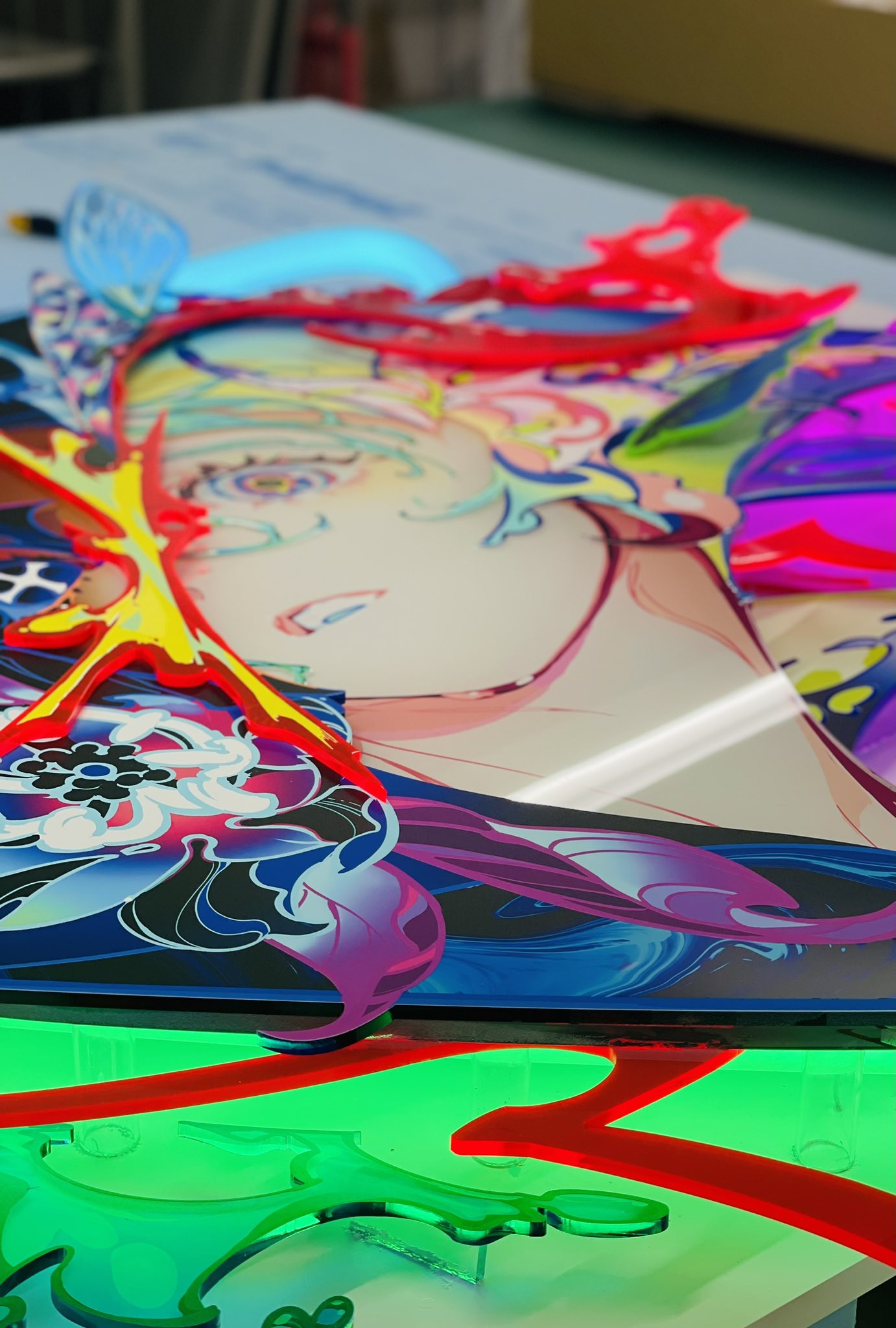 the image showcases a vibrant, multilayered artwork featuring an animestyle woman. crafted using what appears to be acrylic, the piece bursts with neon colors and intricate details, particularly in the character's attire and surrounding floral motifs. the perspective offers a closeup, angled view, highlighting the artwork's depth and the interplay of colors and layers. the bright, artificial lighting further emphasizes the artwork's vividness, creating a captivating and dynamic visual experience.