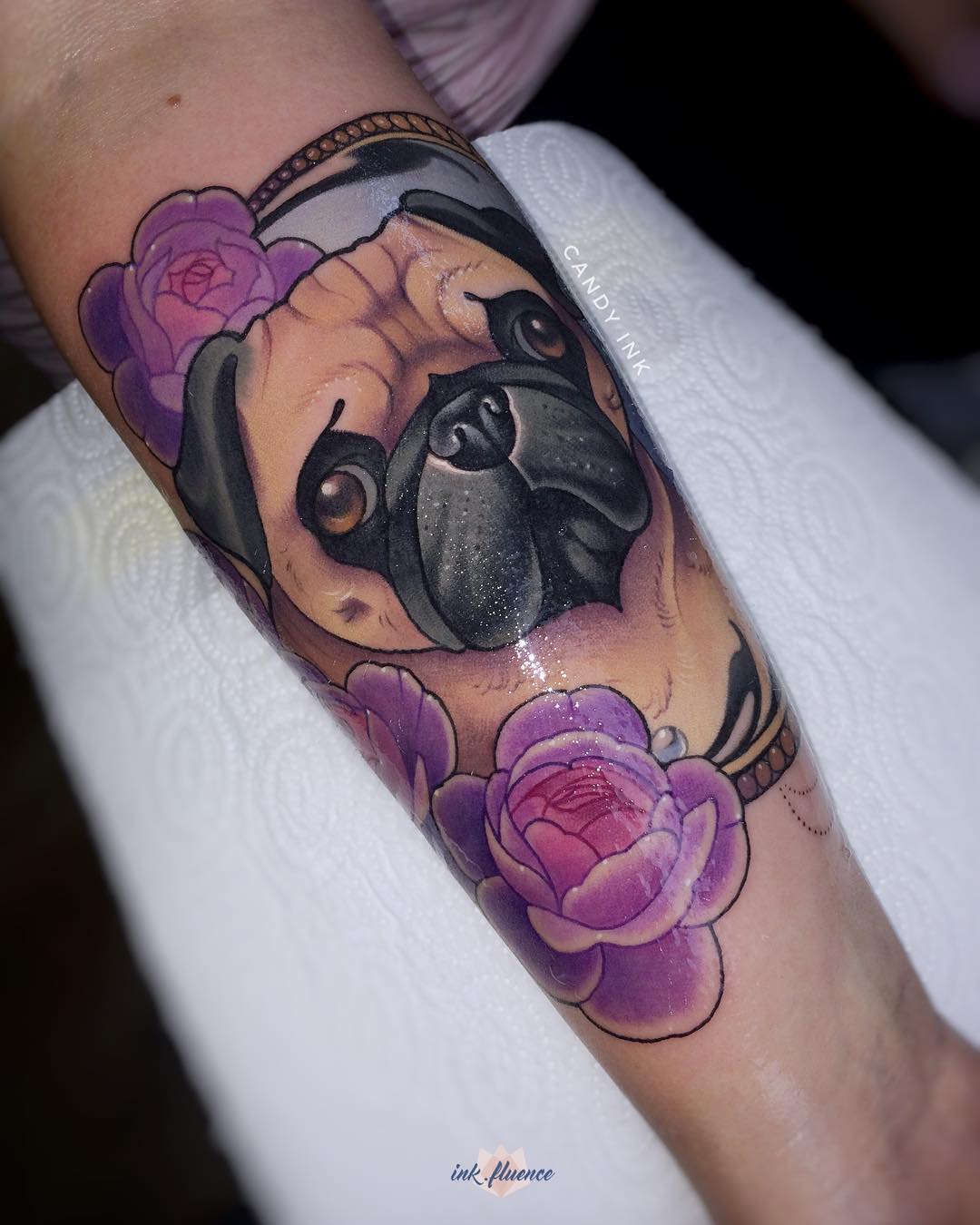 a tattoo of a pug's face surrounded by purple roses on someone's inner forearm. the tattoo is done in a colorful, illustrative style. the tattoo artist's watermark, "candy ink," is visible above the pug's head, and another watermark, "inkfluence," is visible on the bottom right.