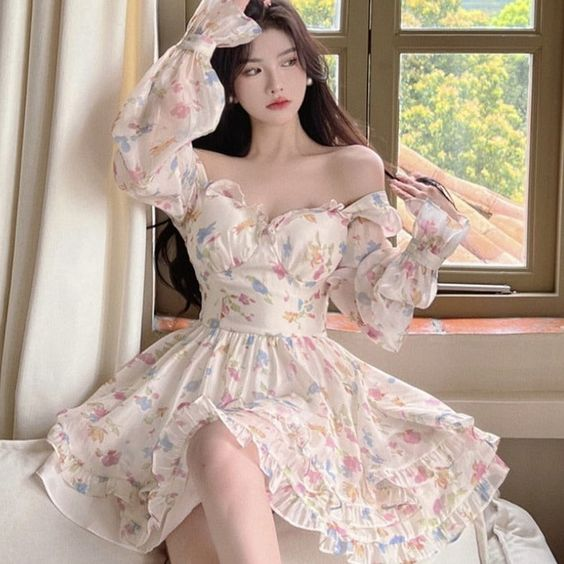 a young woman with long black hair is sitting on a white couch, wearing a white floral dress with an offshoulder neckline and ruffled skirt. the dress has a sweetheart neckline and a fitted bodice. she is posing for the camera with a soft, natural light illuminating her face and the room. behind her is a window with a white frame and a view of greenery. the overall aesthetic of the image is romantic, feminine, and cute.