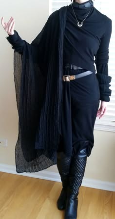 a woman dressed in a black longsleeved dress with a black scarf and black boots. she is looking at the camera, with one hand on her hip and the other arm stretched out. she is wearing a silver necklace and the background is a light brown wall.  the photo was taken at a slight angle.