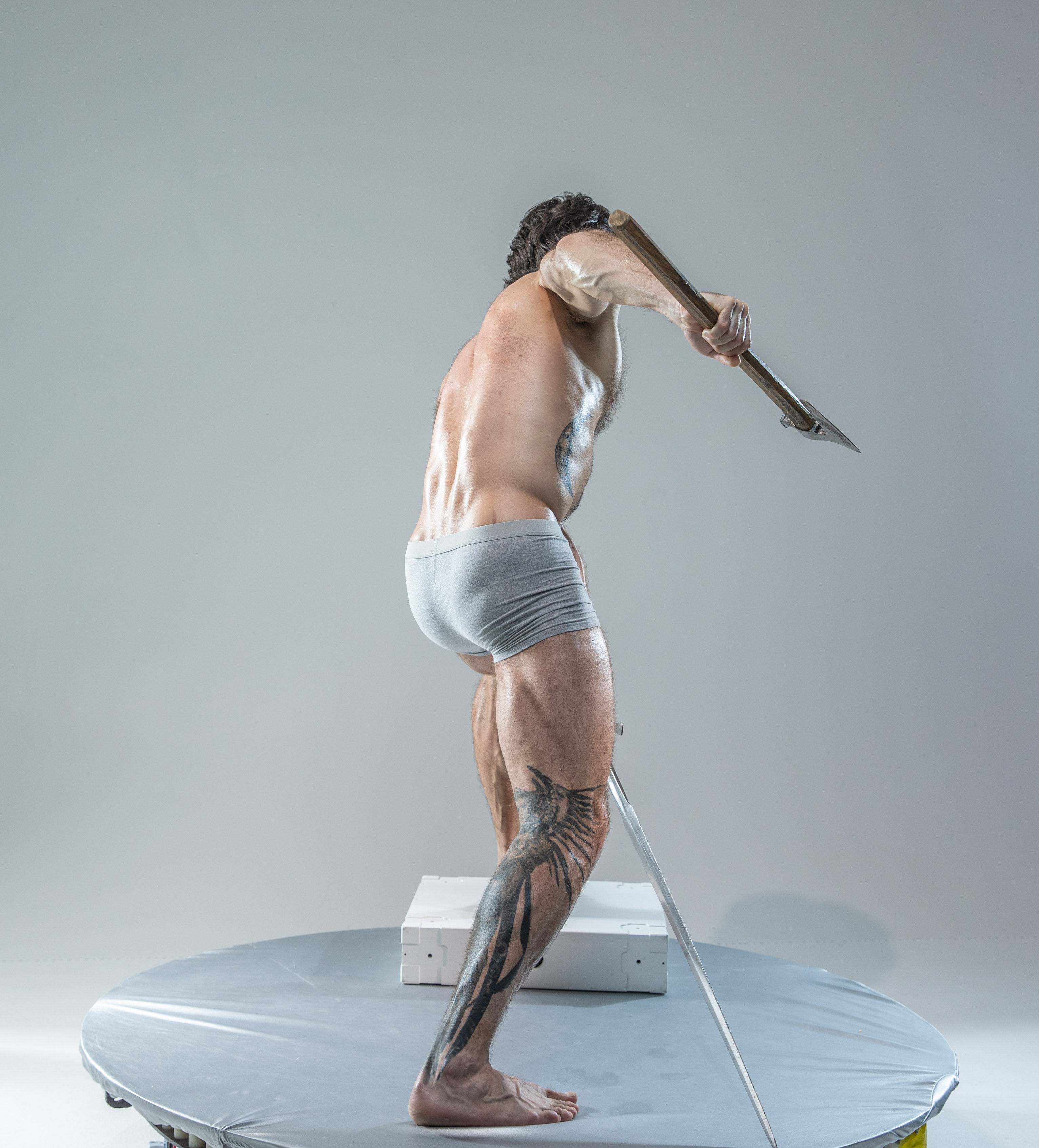 the image showcases a muscular man, captured from behind, midaction as he brandishes a large axe. he stands on a white platform, the soft studio lighting highlighting the definition of his physique. his dynamic pose and the presence of the axe create a sense of power and movement.