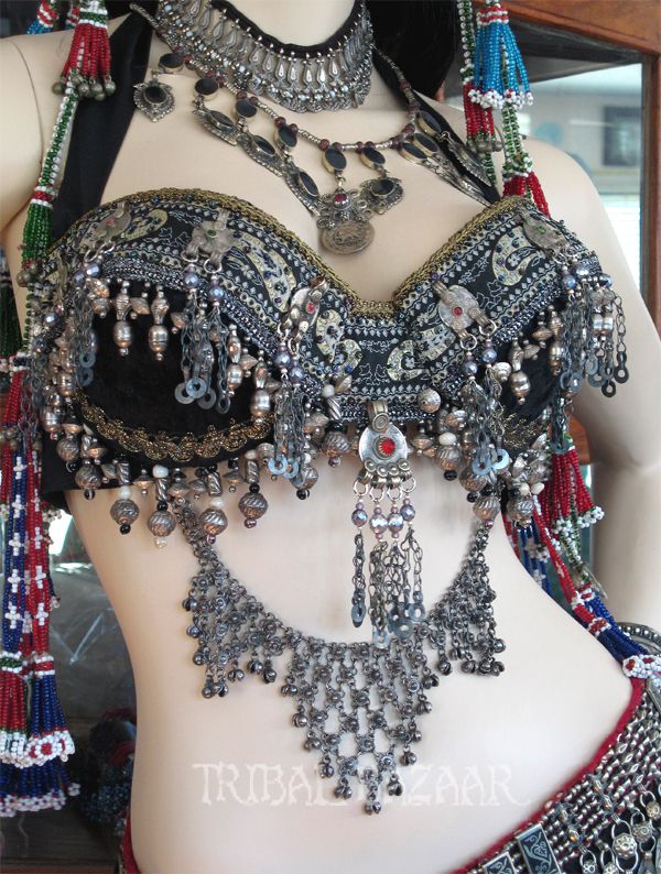 a closeup of a belly dancer wearing an intricate and ornate silver costume.  the costume features a beaded bra and a long chain necklace, both of which are adorned with silver and black beads. the dancer's skin is visible through the costume, and she has a slight smile on her face. the image is taken from a low angle, which emphasizes the beauty of the costume.  the background is blurred, but one can see some red and blue fabrics.
