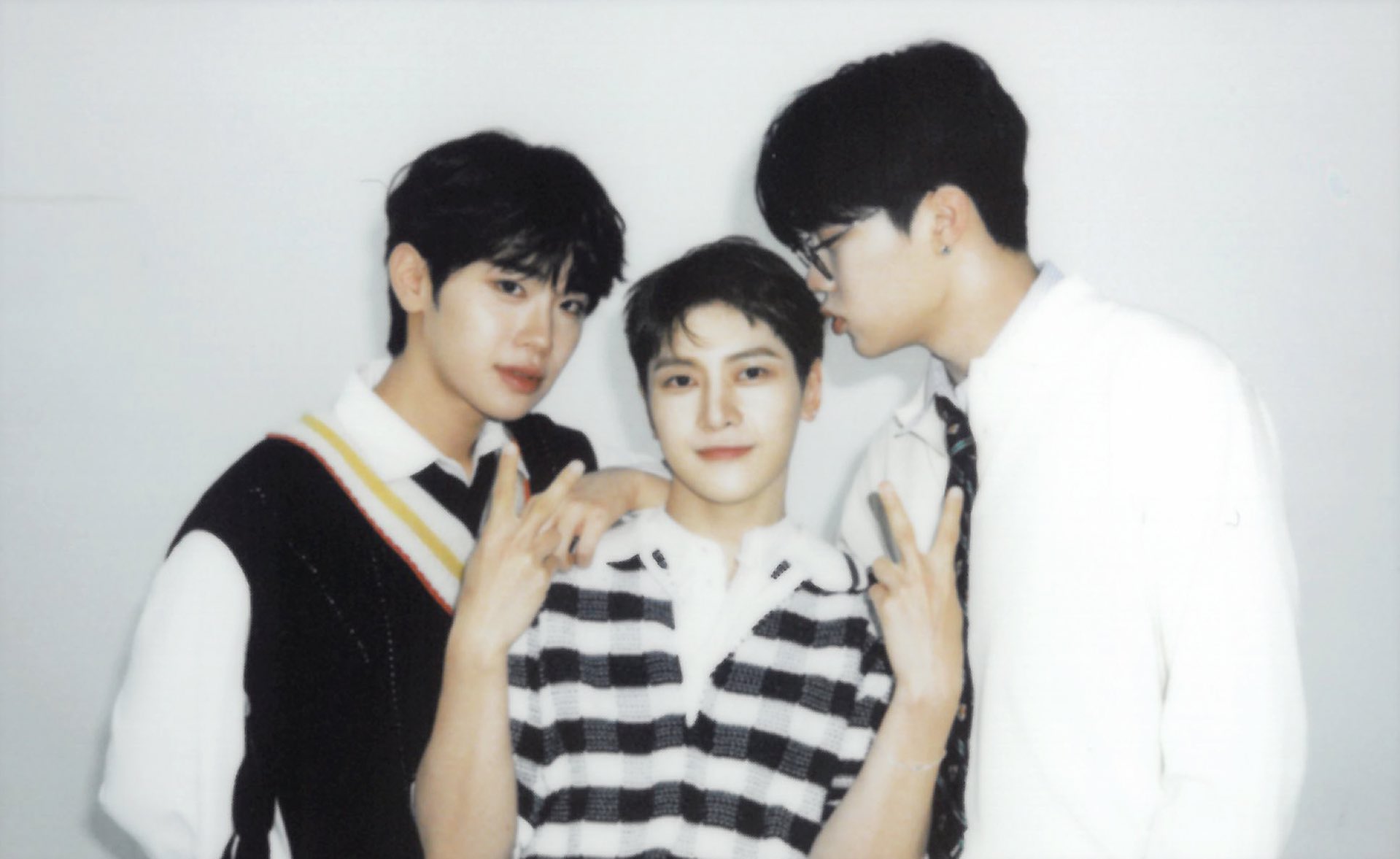 the image is a brightly lit, slightly washedout polaroid selfie of three young men against a plain white background.  they appear to be east asian, possibly kpop idols, and are posing playfully close together. the man on the left wears a black and white striped collared shirt with a black vest featuring diagonal yellow and white stripes. the man in the middle wears a black and white striped collared shirt. the man on the right wears a white collared shirt and black tie. they are all smiling and making peace signs. the overall vibe is friendly, fun, and youthful.