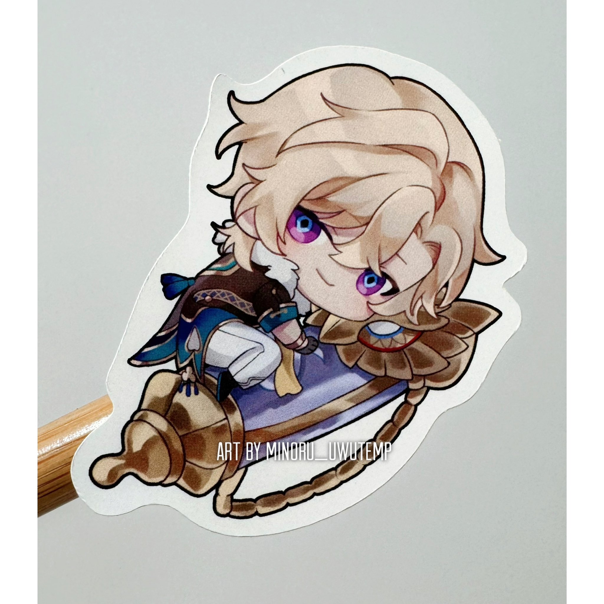 a sticker featuring a chibi anime character, albedo from genshin impact, sitting on a golden scroll. the character has blonde hair, blue eyes, and is dressed in alchemist attire. the background of the sticker is white.