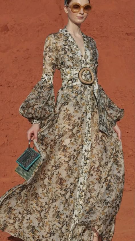 a stylish woman wearing a long floral dress with puff sleeves, a wide belt, and sunglasses. the dress is a mix of beige, brown, and white. the model is posing outdoors against a backdrop of a red brick wall.