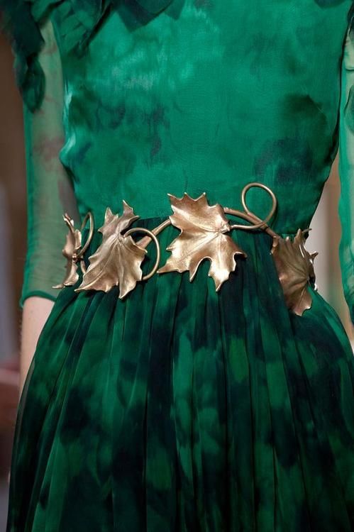 a close up of a woman wearing an emerald green dress with a gold leaf belt. the dress is long and flowing with a loose fit. the belt is made of leaves and is very detailed. the fabric of the dress is soft and flowing and has a subtle pattern. the woman is standing still and the photo is taken from the waist up. the overall tone of the image is very elegant and formal.