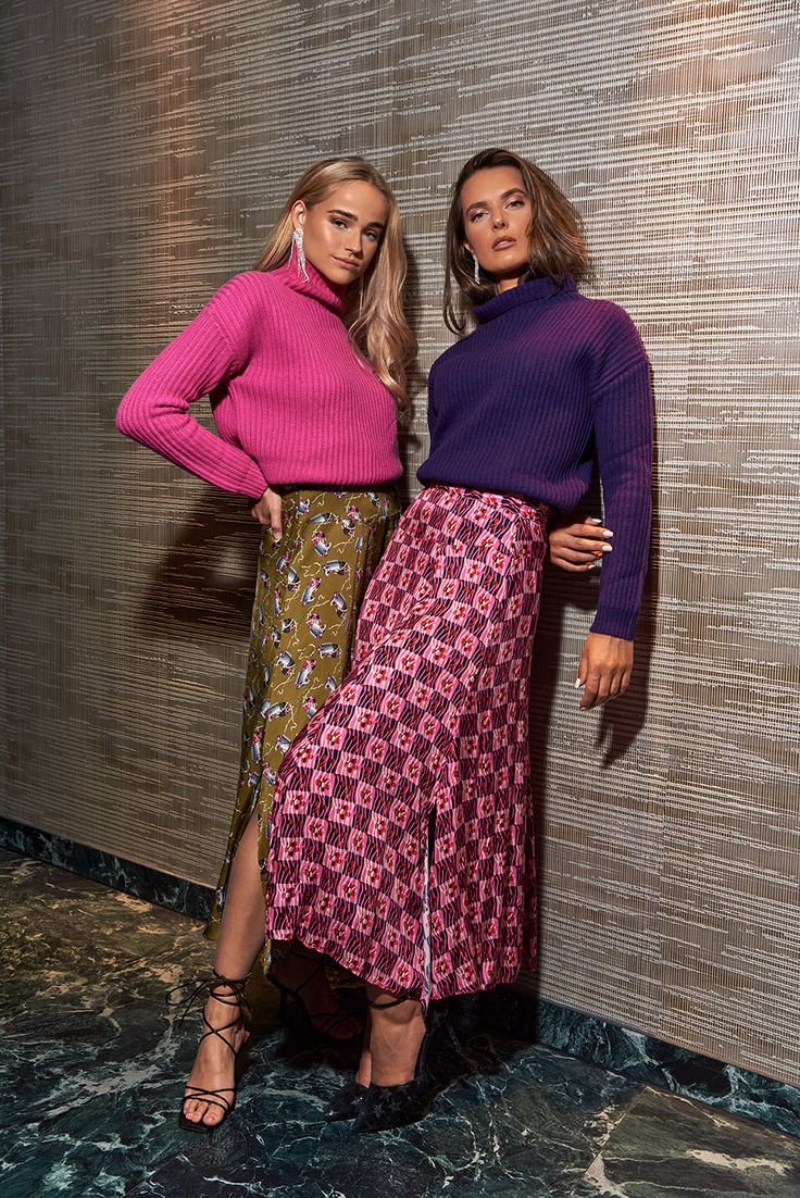 two women are standing close together against a textured wall. one woman is wearing a bright pink turtleneck sweater and a midi skirt with a floral pattern. the other woman wears a deep purple turtleneck sweater and a vibrant pink and maroon checkered midi skirt. both have on dark heels. the scene is indoors, likely in a studio, with marble flooring.  the overall style is elegant and trendy.